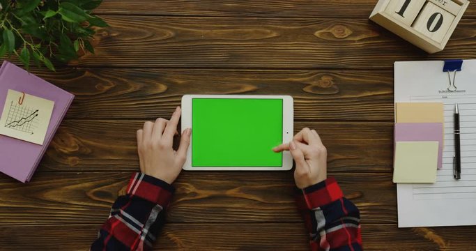 View above on the female hands in the plaid sleeves taping and scrolling on a white tablet device with a green screen horizontally on the wooden desk with office stuff. Chroma key.