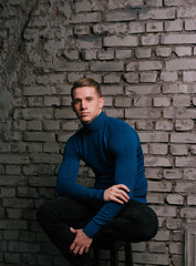 white attractive guy sits on a brick wall background dressed in a blue sweater and black pants