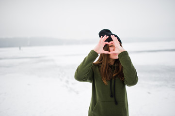 Funny girl wear on long green sweatshirt, jeans and black headwear, at frozen lake in winter day, showing heart on fingers.