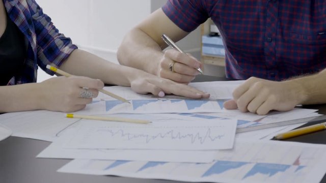 Female and male investors are researching the bitcoin rates in the office, close up. Financial analytics are sitting at the table and building digital currency trends on paper reports with grephs and