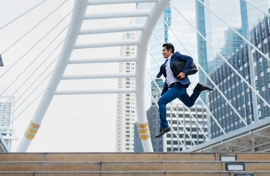 Asian Businessman In Formal Wear Holding Briefcase While Running And Jump In The City On A Background Of Building. Late For Work Concept