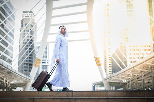 Muslim man wear white turban and dress walking with luggage in public place and modern building background. concept of business travel.