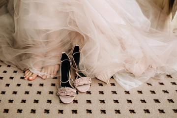Pink elegant shoes of the bride's sandals on the floor next to the feet and a wedding gentle dress made of translucent fabric
