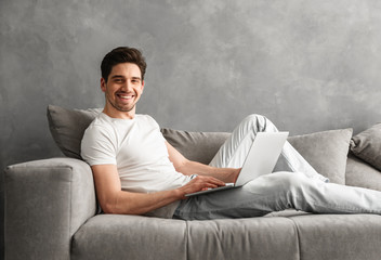 Caucasian unshaved man 30s in casual clothing using notebook, while lying on cozy sofa in gray...