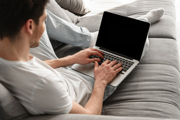 Photo from top of young brunette guy 30s in casual jeans working on notebook, while lying on cozy gray sofa at home