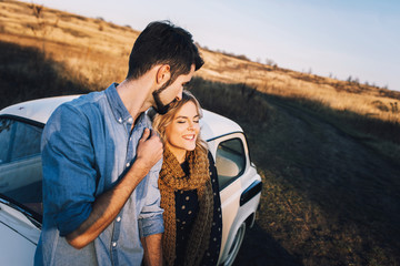 Young beautiful couple hugging each other, sitting on a small white car in beautiful evening light. Stylish guy with a beard and blond girl laughing