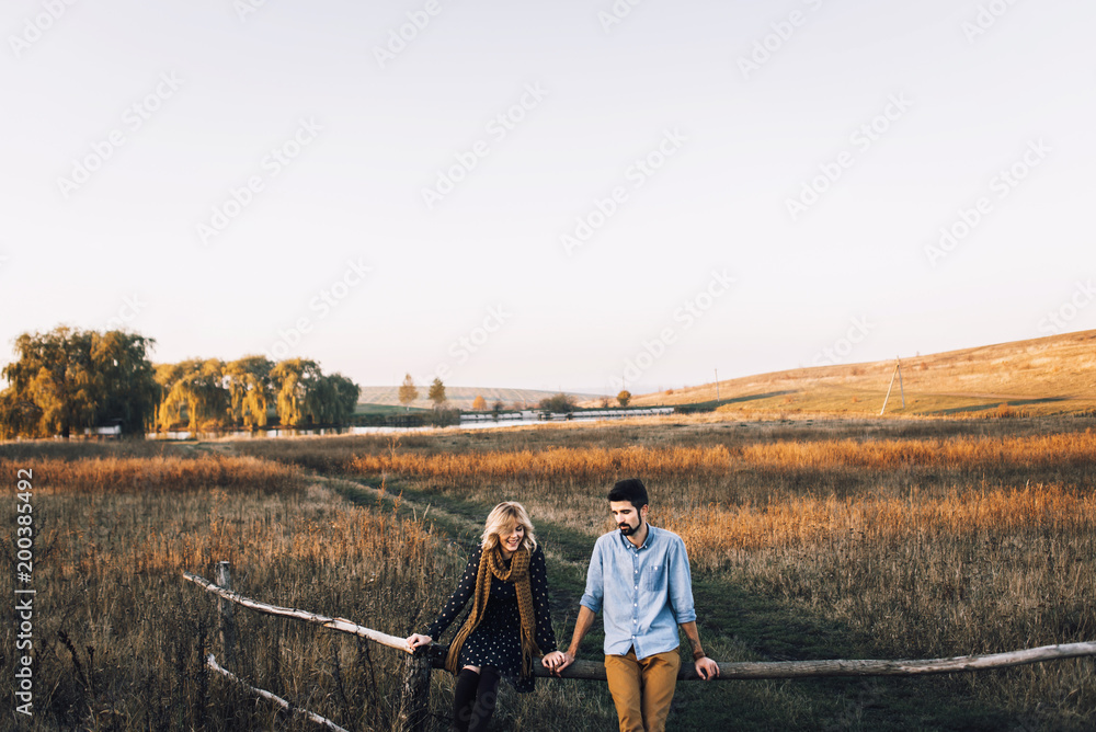 Wall mural handsome guy with a beard in a denim shirt hugs and holds the hand of a beautiful girl in a blue dress and yellow scarf in a field at sunset. stylish couple sitting on a wooden fence and look around