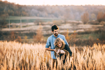 handsome guy with a beard in a blue denim shirt gentle hugs, hand holding and kissing a girl with blond hair in a blue dress and yellow scarf in a field at sunset. stylish couple