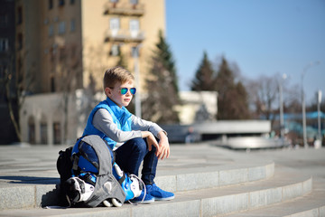 Young boy sitting near grey wall with inline roller skates and all protection at outdoor skate park
