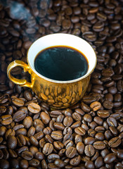 Fresh hot coffee steaming out of an ornate gold tea cup on a pile of coffee beans