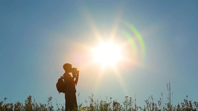 Boy walks along grassy hill using old vintage camera for filming video. Kid's silhouette isolated at bright blue sky and sunshine background. 