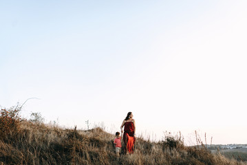 beautiful young mother in a long red dress easy walks with her young son. Summer, sunset, high yellow grass, mountains. He is on the hands, holding the hand