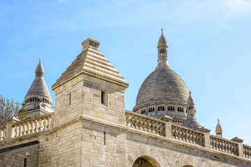Fototapeta premium View of the dome and steeple of the Basilica of the Sacred Heart of Paris with the outer stone wall and a turret of the great water reservoir of Montmartre in the foreground under a clear blue sky.