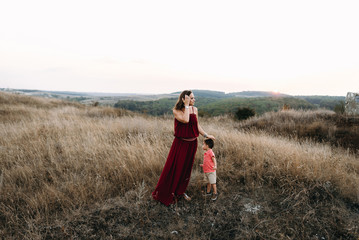 beautiful young mother in a long red dress easy walks with her young son. Summer, sunset, high yellow grass, mountains. He is on the hands, holding the hand