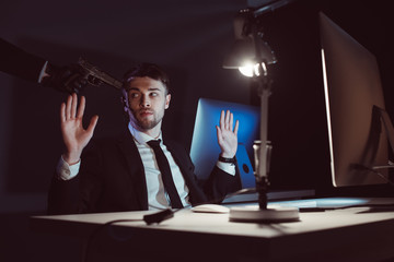 agent with gun at head and hands up sitting at table with computer screen in dark