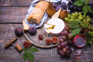 Red wine in a glass,  wine bottle, cork, bottle screw and a set of products - cheese, grapes, nuts, pears, plums, flowers, figs, white bread, baguette on a wooden board, background