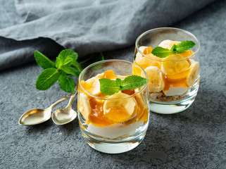 Healthy layered dessert with yogurt, banana, mango jam, cookie in glass on gray stone background, side view.