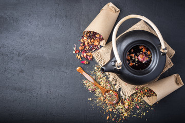 Black ceramic teapot with tea,Different tea and herbs , dried rose flowers on dark concrete background.Top view,copy space.