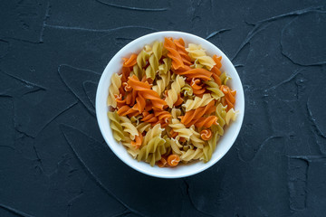 Multicolor spiral macaroni Pasta in a white bowl on a black textured background from the side.