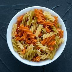 Multicolor spiral macaroni Pasta in a white bowl on a black textured background from the side. Close-up with the top.
