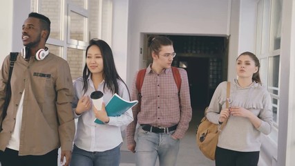 Four groupmates of different ethnicity are walking in long spacious wide glassy corridor in university smiling and laughing and talking to each other