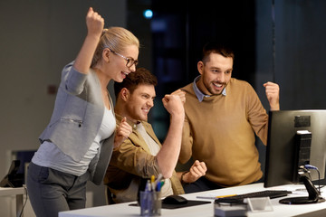 business, success and teamwork concept - happy coworkers with computer celebrating victory at night office