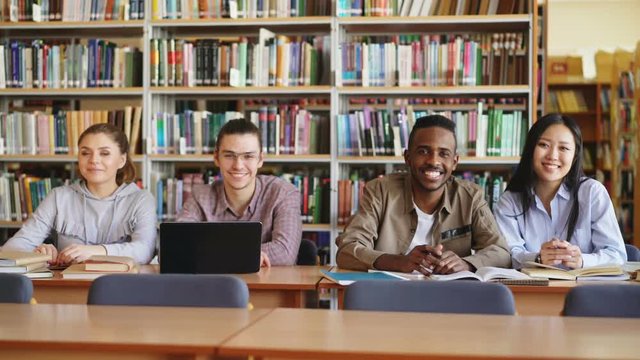 Portrait of four multi-ethnic students sitting at long desk in big spacious library with piles of books looking at camera and smiling positively