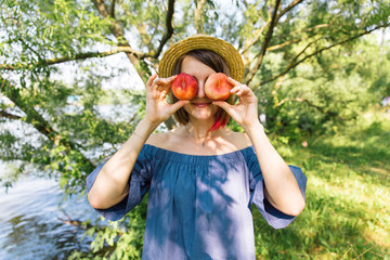 Happy woman in straw hat covering her eyes with peaches on garden background. Summer, outdoors. Close up portrait of young woman holding red peaches instead of her eyes. Organic food, fruit diet.