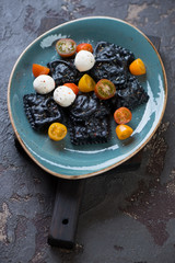 Turquoise plate with black ravioli, tomatoes and mozzarella on a brown stone background, studio shot