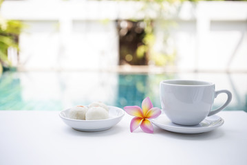 Fototapeta na wymiar Afternoon tea time at pool side, white tea cup with thai sweet on white table over blurred swimming pool background, summer outdoor day light