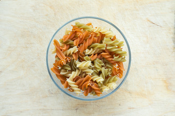 Multicolor spiral macaroni pasta in a glass bowl on a wooden table texture background, in the center close-up with the top.