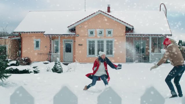 Young Beautiful Couple Throws Snowballs at Each other While Snow Falls. Happy Man and Woman Playing with Snow in the Yard of their Idyllic House. Shot on RED EPIC-W 8K Helium Cinema Camera.