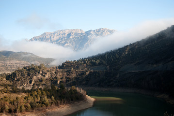 Embalse con  paisaje de montaña y bancos de niebla al fondo.