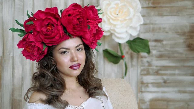 Girl posing in front of camera. young woman in a wreath of scarlet peonies on her head, dark long curly hair descends on the shallow shoulders.