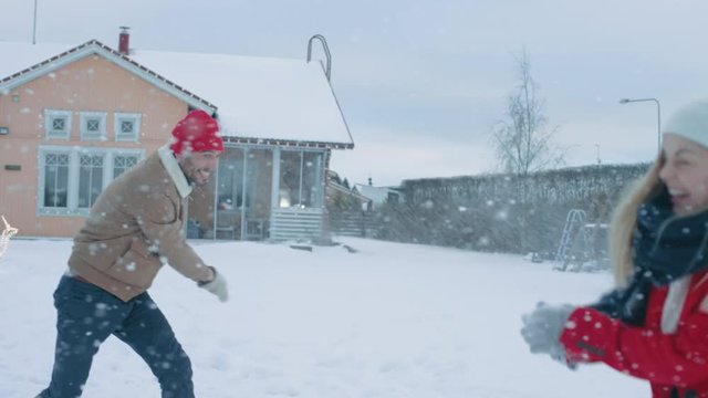 Young Beautiful Couple Throws Snowballs at Each other While Snow Falls. Happy Man and Woman Playing with Snow in the Yard of their Idyllic House. Shot on RED EPIC-W 8K Helium Cinema Camera.