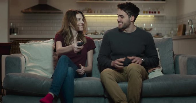Young, attractive couple sit down on a couch and turn on the tv using the remote control.