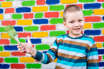Happy charming Caucasian child painting wooden boat against brick wall background. Brush, container with paint. Romantic sunset and lighting. Preparation of background and props for photography.