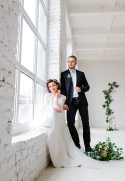 Wonderful wedding couple posing in a bright white room with large windows.