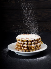White plate with homemade Belgian waffles, on top of poured sifting of powdered sugar on black background, very tasty snack. sugar over old wooden table. Dark rustic style. copy space, stylish
