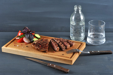 On a light wooden board juicy steak cooked on the grill. Next to the steak is a salad of fresh vegetables. In the frame of the dining room, appliances, a glass and a bottle of water. Close-up.
