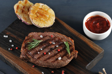 On a wooden board, beef steak cooked on the grill. Next to the steak are two halves of garlic, cooked on the grill. In the frame a cup with ketchup. View from above. Macro photography.