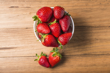White bowl with some strawberries on a wooden table; two strawberries are out of the bowl on the table.