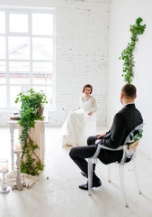 A young couple in a wedding dress sitting on chairs in a white studio.