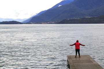 Fototapeta na wymiar Mann steht auf einem Steg, Mit Jogginghose und Kaputzenshirt begleitet, am Comer See mit ausgebreiteten Armen und blickt nach Colico
