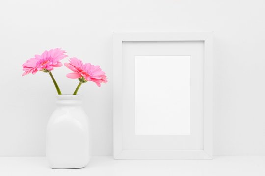 Mock up white frame and pink daisy flowers in vase on a shelf or desk. White color scheme. Portrait frame orientation.