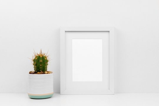 Mock up white frame and cactus in pot on a shelf or desk. White color scheme. Portrait frame orientation.