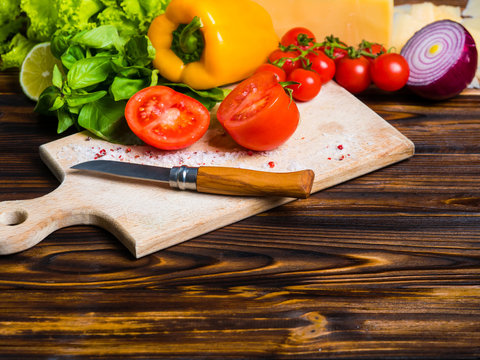 Appetizing still life. Fresh bulgarian yellow pepper, cherry tomatoes, cheese, lettuce leaves, red onion, green basil on wooden board. The knife for cutting vegetables. Top view - Powered by Adobe