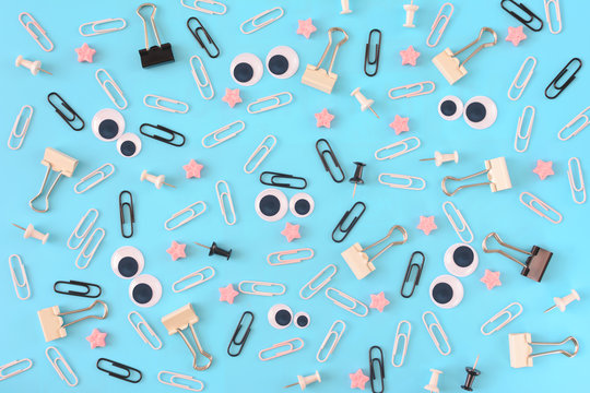 White and black paper clips, pink stars and stationery are scattered chaotically on a blue background. Funny smiley from clips in the center. Photo from the top.
