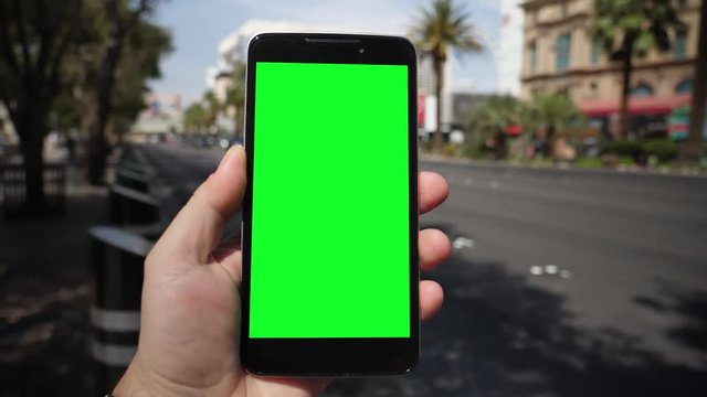 A man holds a smartphone outside near Las Vegas Boulevard as traffic and taxis pass by in the background. Green screen with optional corner pin markers for advanced screen replacement.  	