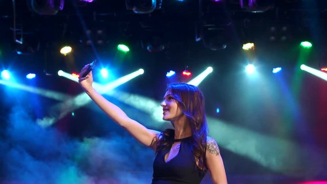 Portrait of happy girl in evening dress with a telephone on a party. Funny girl dancing in a nightclub and makes a selfie. Slow motion. Dark. Multi-colored spotlights.